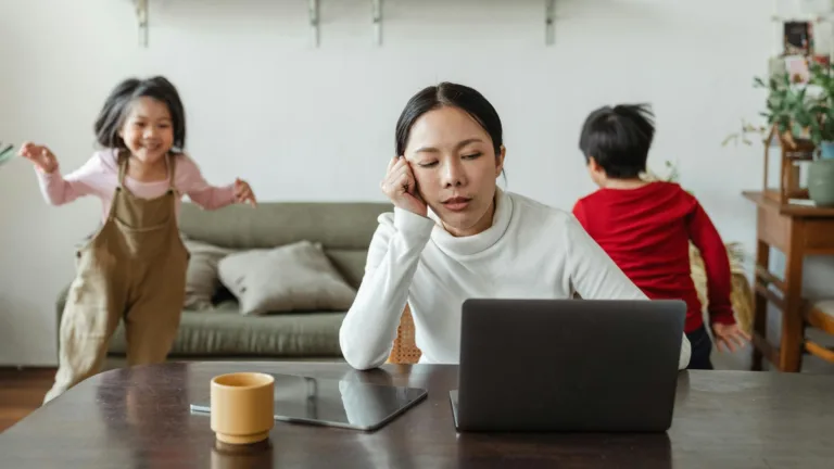 Stressed mother working at laptop while children play in the background, symbolizing anxiety and emotional overload