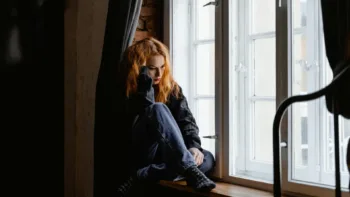 Young woman sitting alone by a window, looking down in sadness — symbolizing the emotional weight of depression before treatment