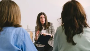 Therapist speaking with two clients in a small group setting, holding a clipboard during a focused session.