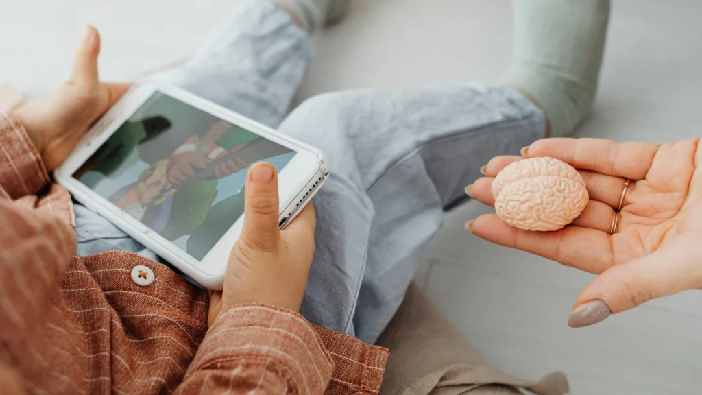 Close-up of a hand offering a brain model to a child using a tablet, representing education about the brain and central nervous system