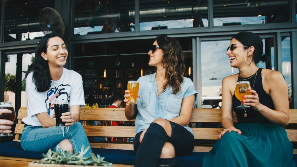 Three women laughing and drinking together outside a bar on a sunny day
