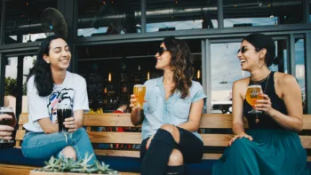 Three women laughing and drinking together outside a bar on a sunny day