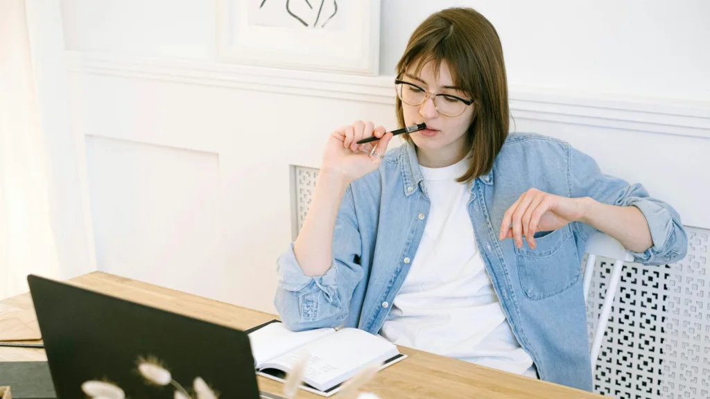 Woman sitting at desk looking thoughtfully at laptop, holding a pen and open notebook