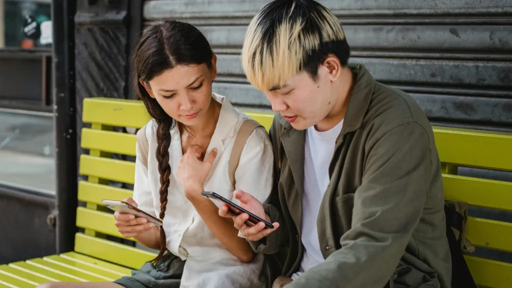 Couple sitting on a bench looking at their phones with distracted expressions, suggesting relationship tension