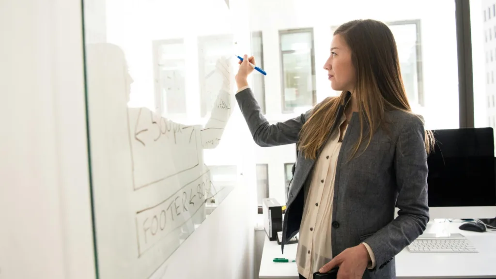 Female educator writing on a whiteboard in a bright classroom, symbolizing the professional and personal mental health journey of public school teachers