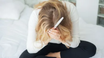 Woman sitting on a bed, holding a pregnancy test and crying with her head in her hands, overwhelmed by infertility struggles