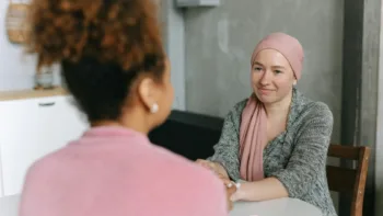 Woman wearing a headscarf holding hands across a table with a friend, symbolizing emotional support during cancer treatment.