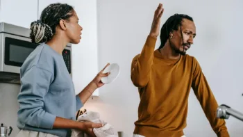 Frustrated couple in kitchen having a disagreement, with one partner drying dishes and the other raising a hand dismissively.
