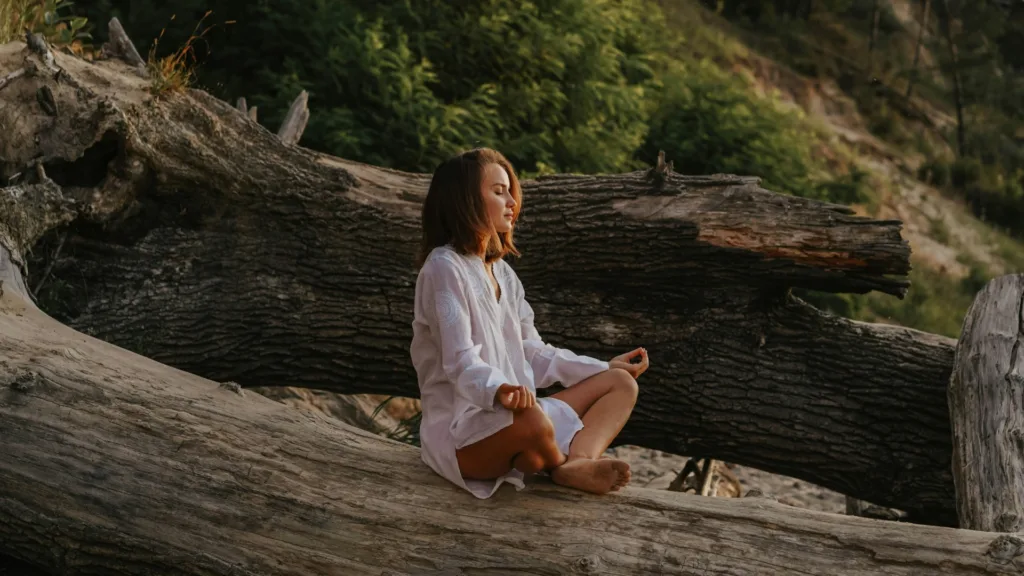 Woman sitting cross-legged on a fallen tree with eyes closed, practicing mindfulness outdoors in nature