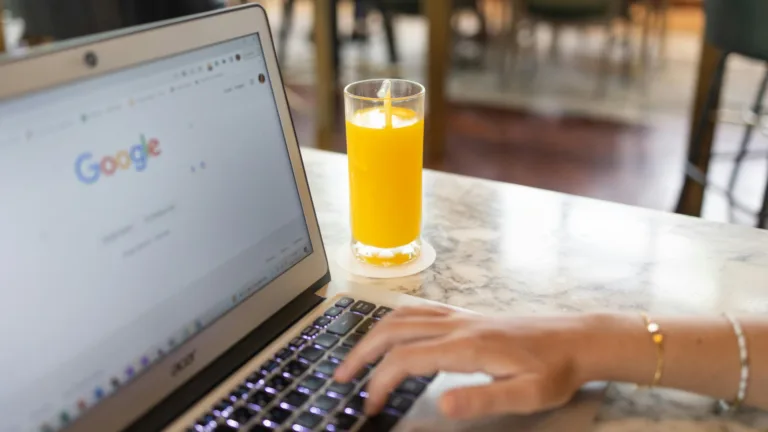 Person using laptop with Google search open, next to a glass of orange juice on a marble table while searching google