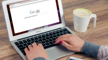 Close-up of hands typing on a laptop with Google search open, next to a coffee mug on a wooden table