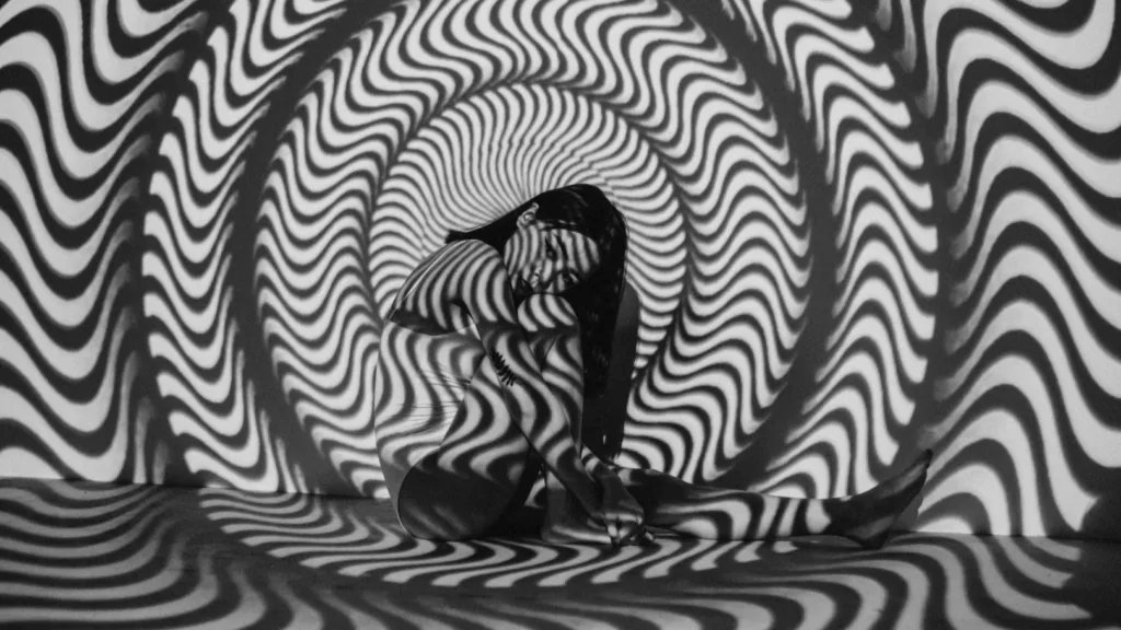 Person seated in a fetal position surrounded by swirling black-and-white hypnotic patterns, symbolizing emotional overwhelm or altered states