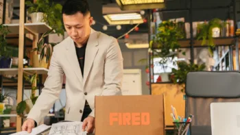 Man packing personal items into a box labeled “Fired” after a layoff in a modern office