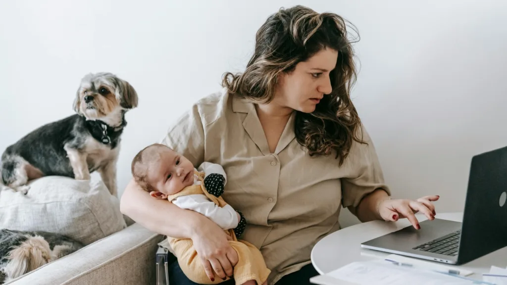 Stressed mother multitasking with a baby in her arms while working on a laptop, with a dog beside her, illustrating the emotional toll of doing too much