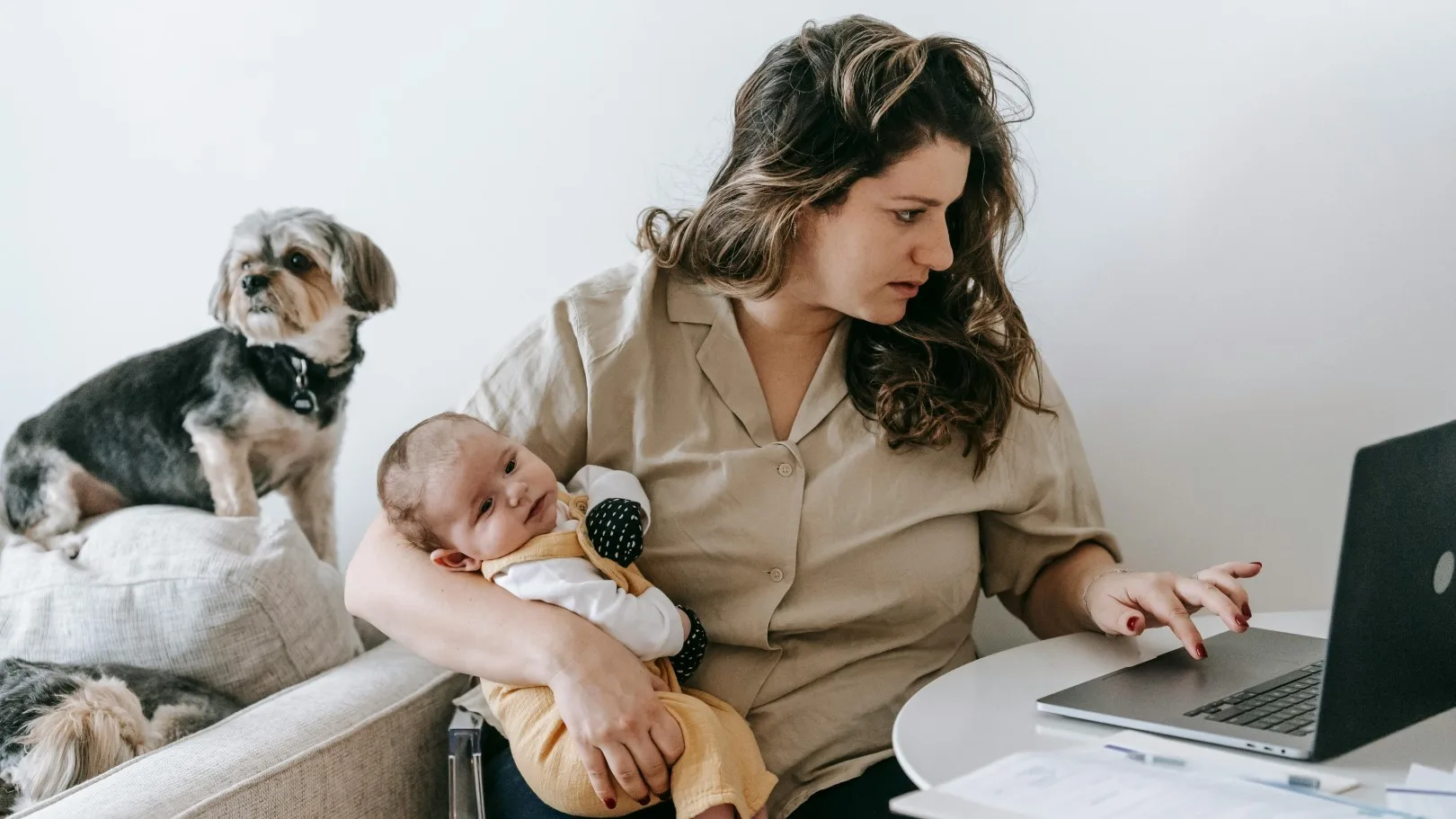 Stressed mother multitasking with a baby in her arms while working on a laptop, with a dog beside her, illustrating the emotional toll of doing too much