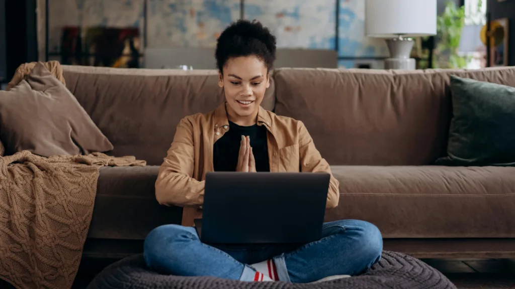 Smiling woman sitting cross-legged in front of laptop, expressing gratitude or connection during an online therapy session from home