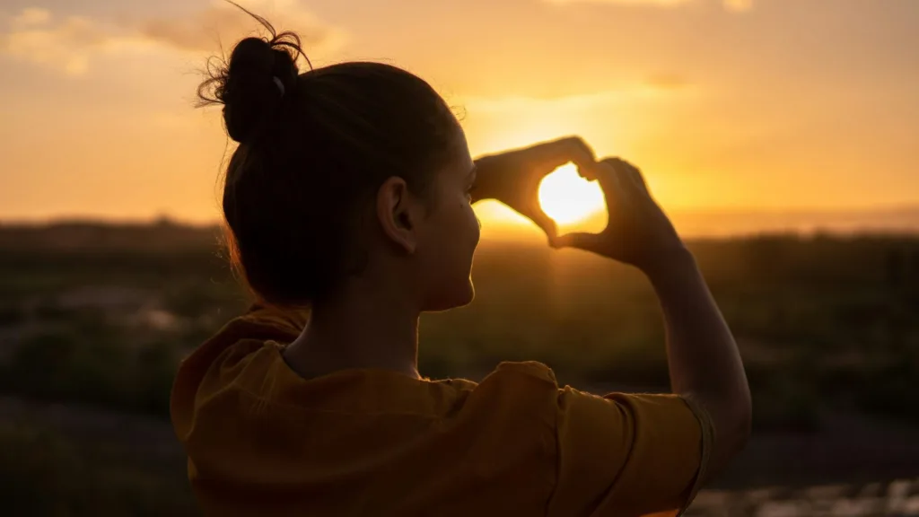 Woman forming a heart shape with her hands at sunset, symbolizing self-compassion
