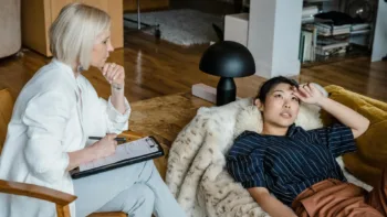 Therapist taking notes while woman lies back on couch, appearing emotionally exhausted during a counseling session