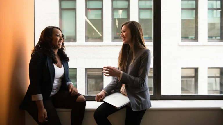 Two female therapists sitting by a window, smiling and collaborating with laptop and notes in hand