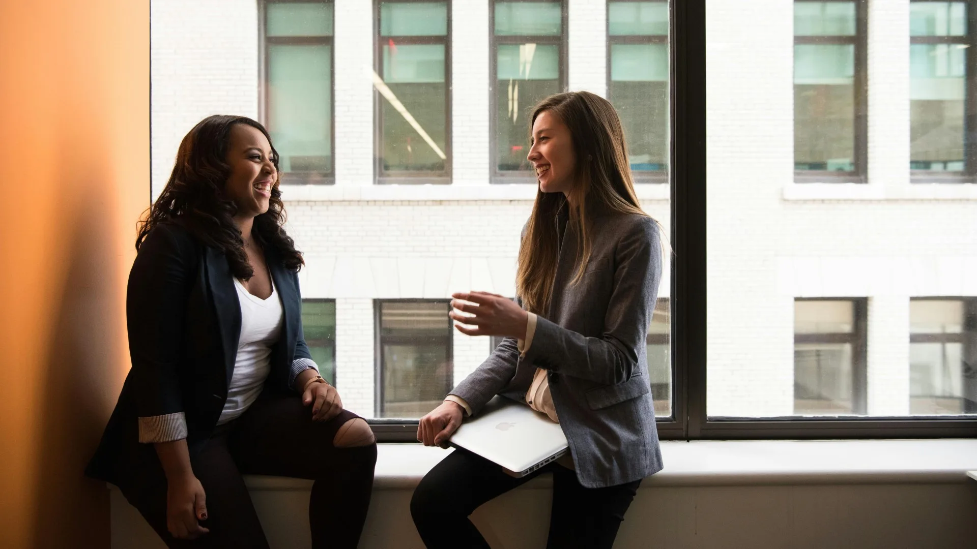 Two female therapists sitting by a window, smiling and collaborating with laptop and notes in hand